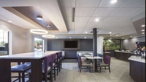 A modern restaurant with high tables and chairs, a large TV screen, and a kitchen area. - La Quinta Inn & Suites by Wyndham Jackson Airport/Pearl (Pearl)