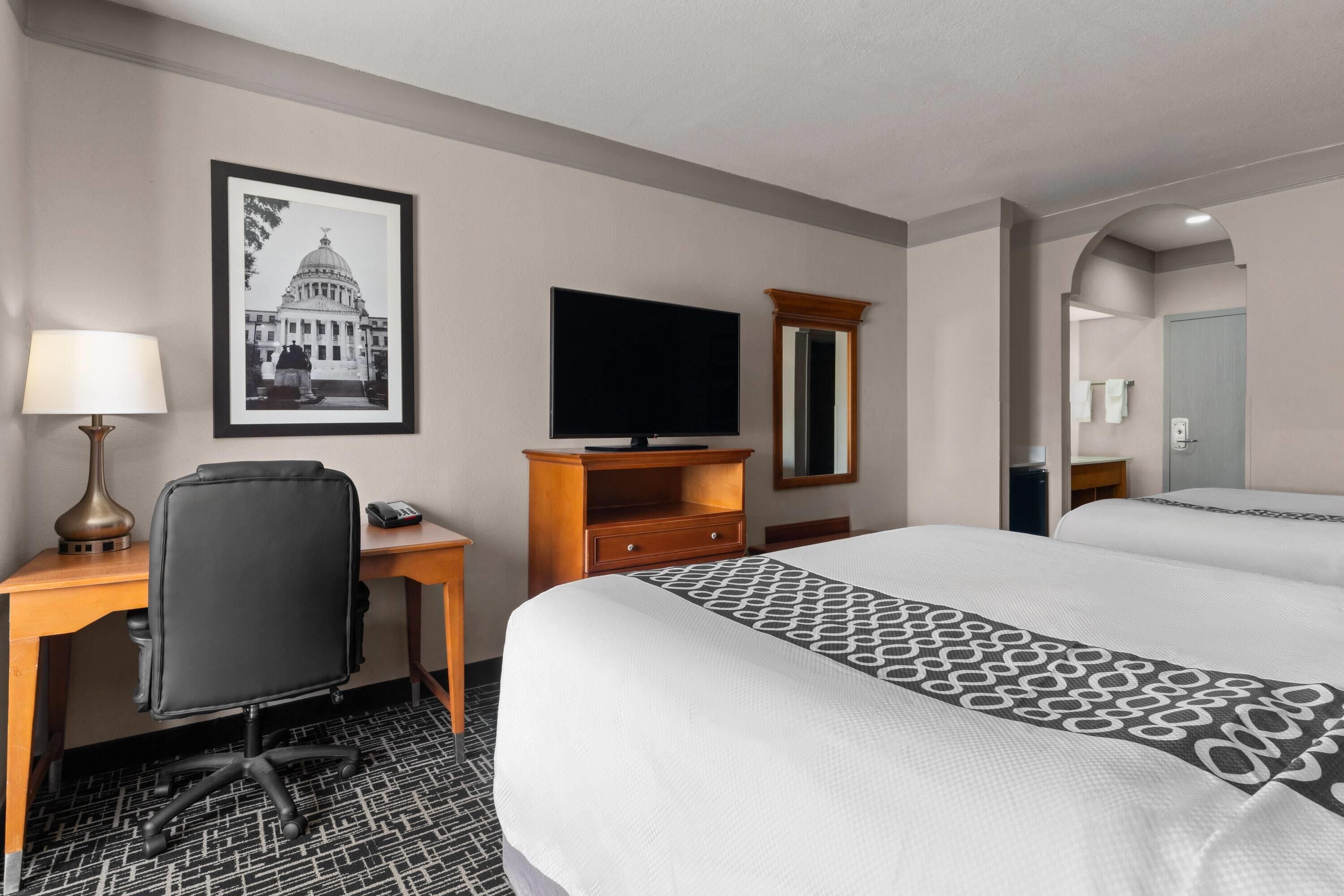 Una habitación de hotel con una cama, un escritorio con lámpara, una televisión, un espejo y un cuadro en la pared.