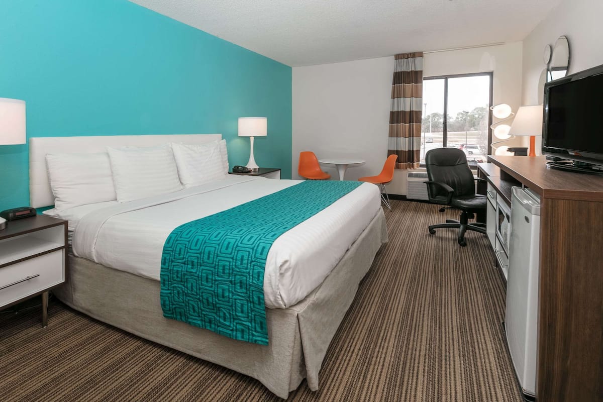 A hotel room with a bed, a desk with a television, two chairs, a desk lamp, and a window with curtains.