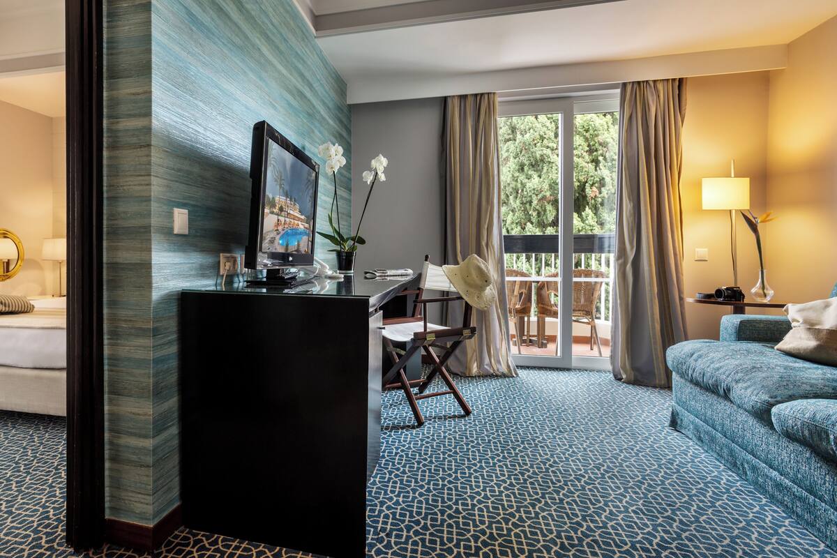 Ein Hotelzimmer mit einem großen Bett, einem Schreibtisch mit Computer, einem Sessel und einem Balkon mit Blick auf Bäume.