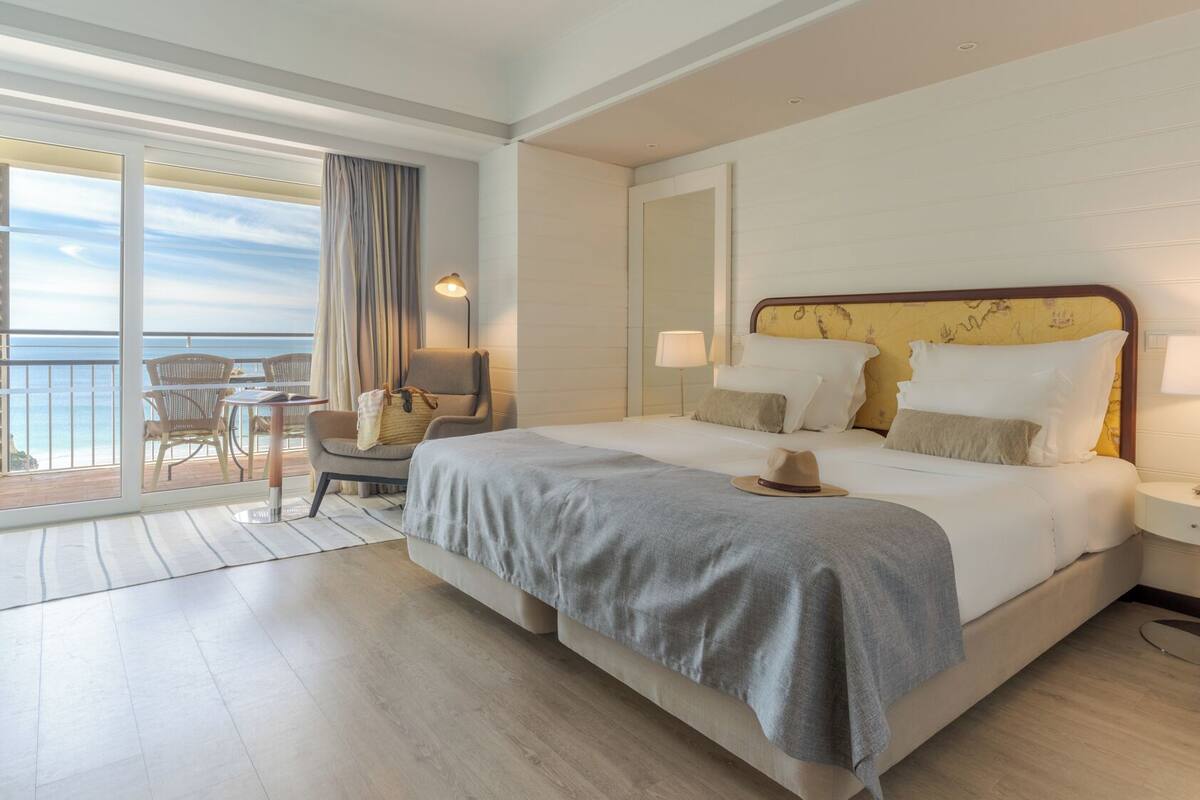 Ein Hotelzimmer mit einem großen Bett, zwei Nachttischen, einem Sessel, einem Balkon mit Meerblick und einem großen Fenster mit Vorhängen.