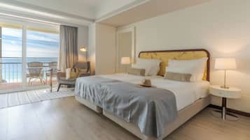 A hotel room with a large bed, two bedside tables, a chair, a balcony with a view of the sea, and a large window with curtains.