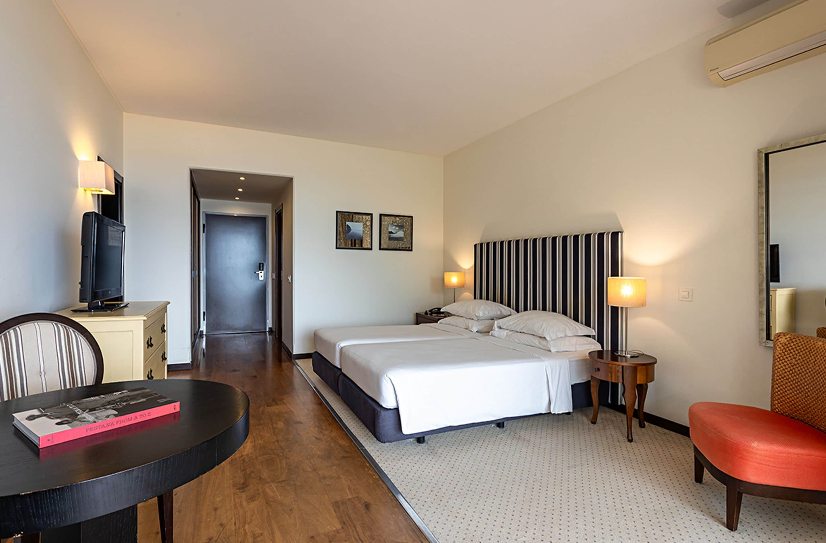 Una habitación de hotel con cama, mesitas de noche, un escritorio con silla, televisor y una mesa de comedor.