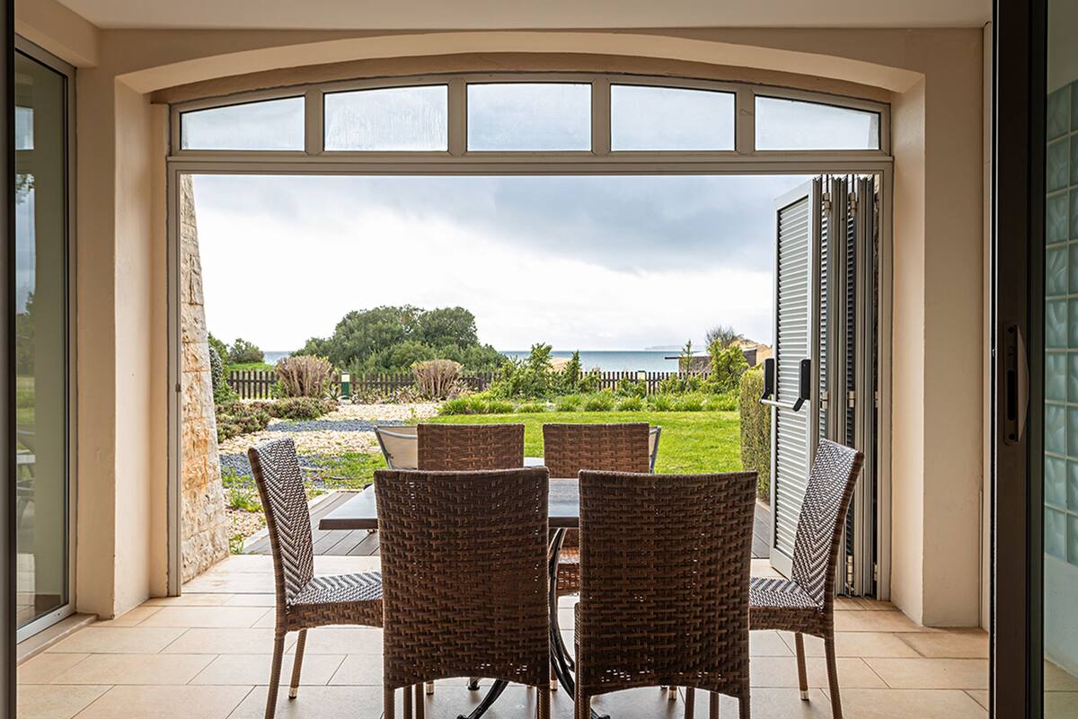 Eine offene Terrasse mit Tisch und Stühlen, mit Blick auf einen Garten und das Meer durch große Fenster.