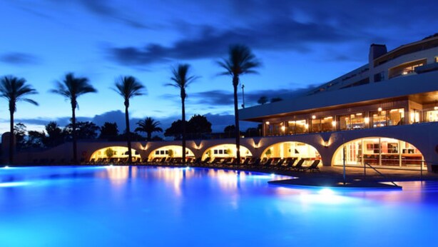Una vista notturna di un albergo con piscina, palme e un edificio illuminato con strutture ad arco.