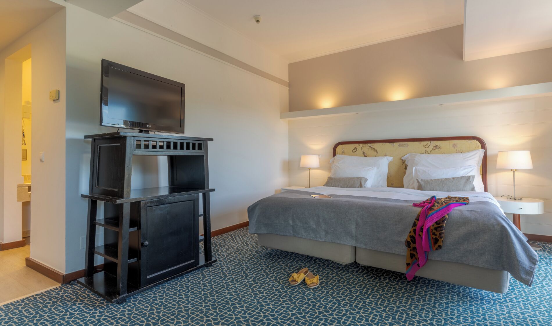 Una habitación de hotel con cama, lámparas de noche, un televisor sobre un soporte y un piso con diseño.