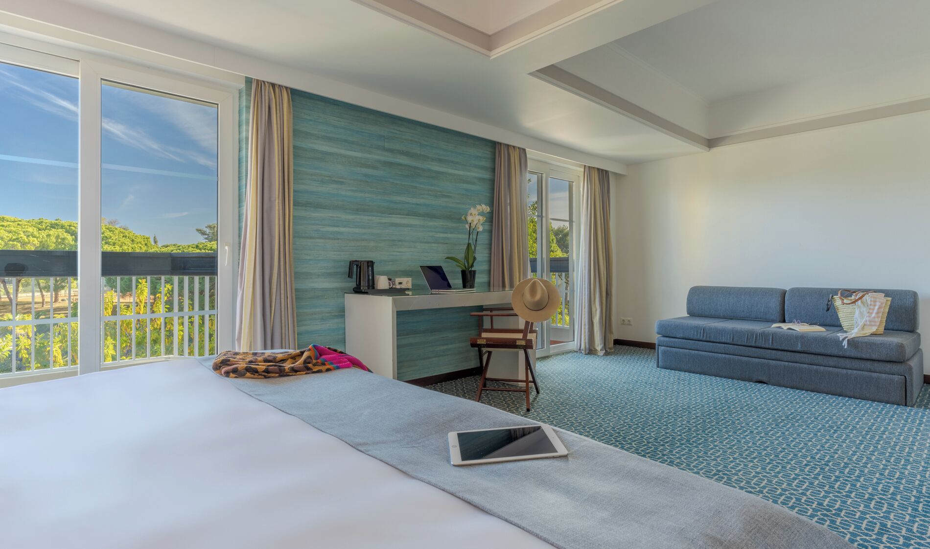 Habitación de hotel con cama, escritorio con computadora portátil, sofá y balcón con vista a la vegetación.