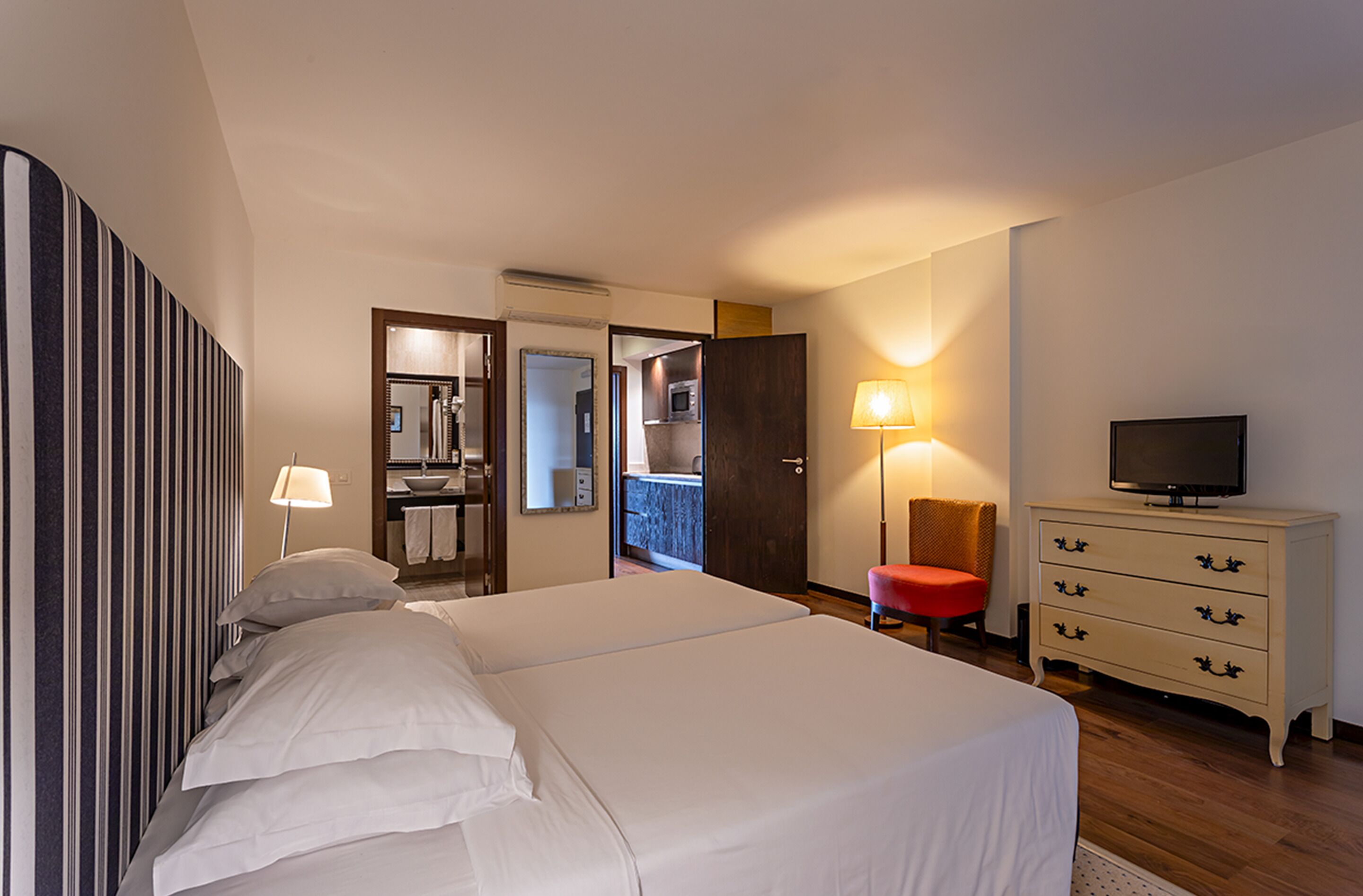 A hotel room with a large bed, a television, a dresser, a chair, and a bathroom visible through an open door.