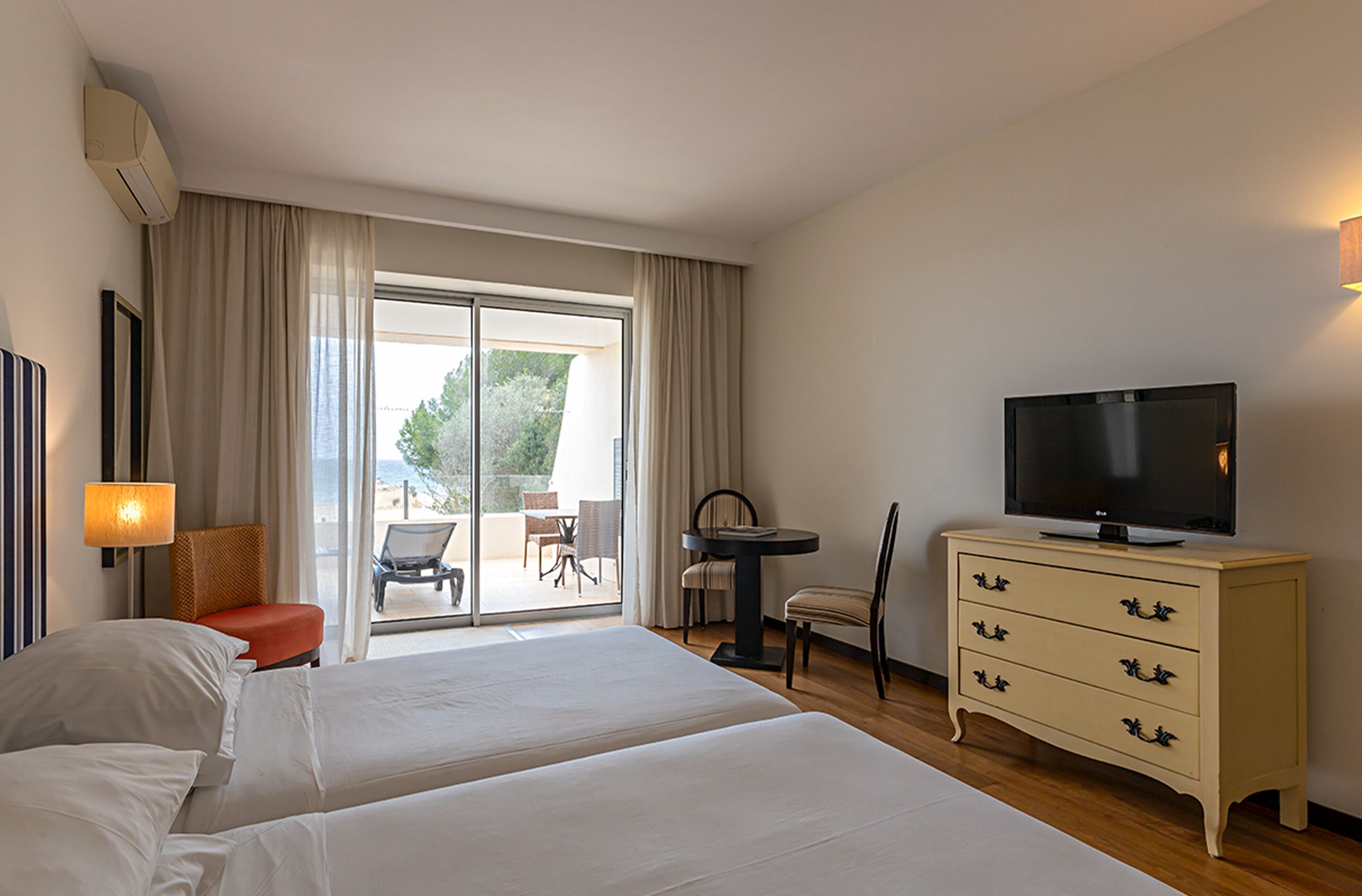 A hotel room with two beds, a TV, a dresser, and a balcony with outdoor seating.