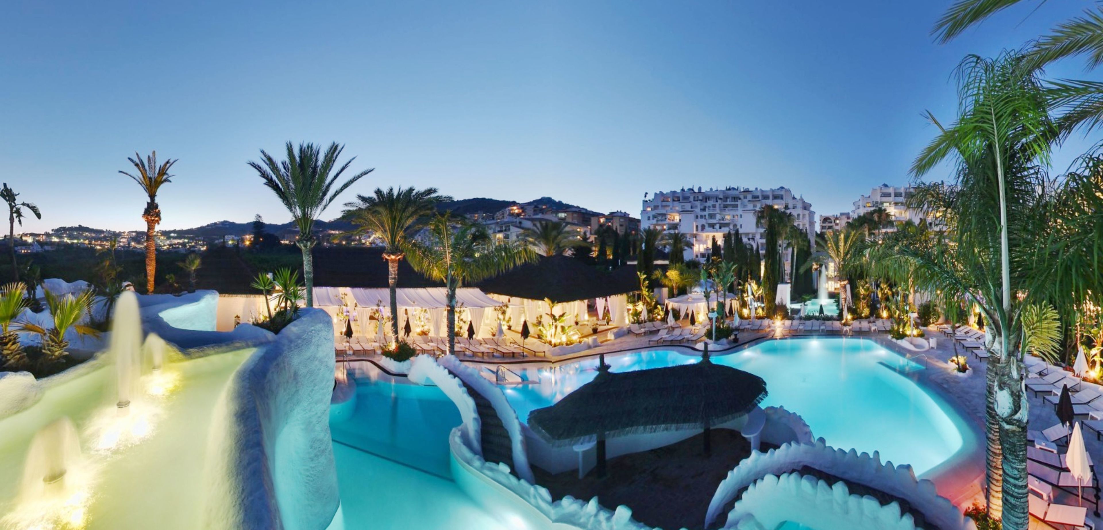 Un complexe hôtelier doté d’une grande piscine, de palmiers et surplombant un paysage montagneux.