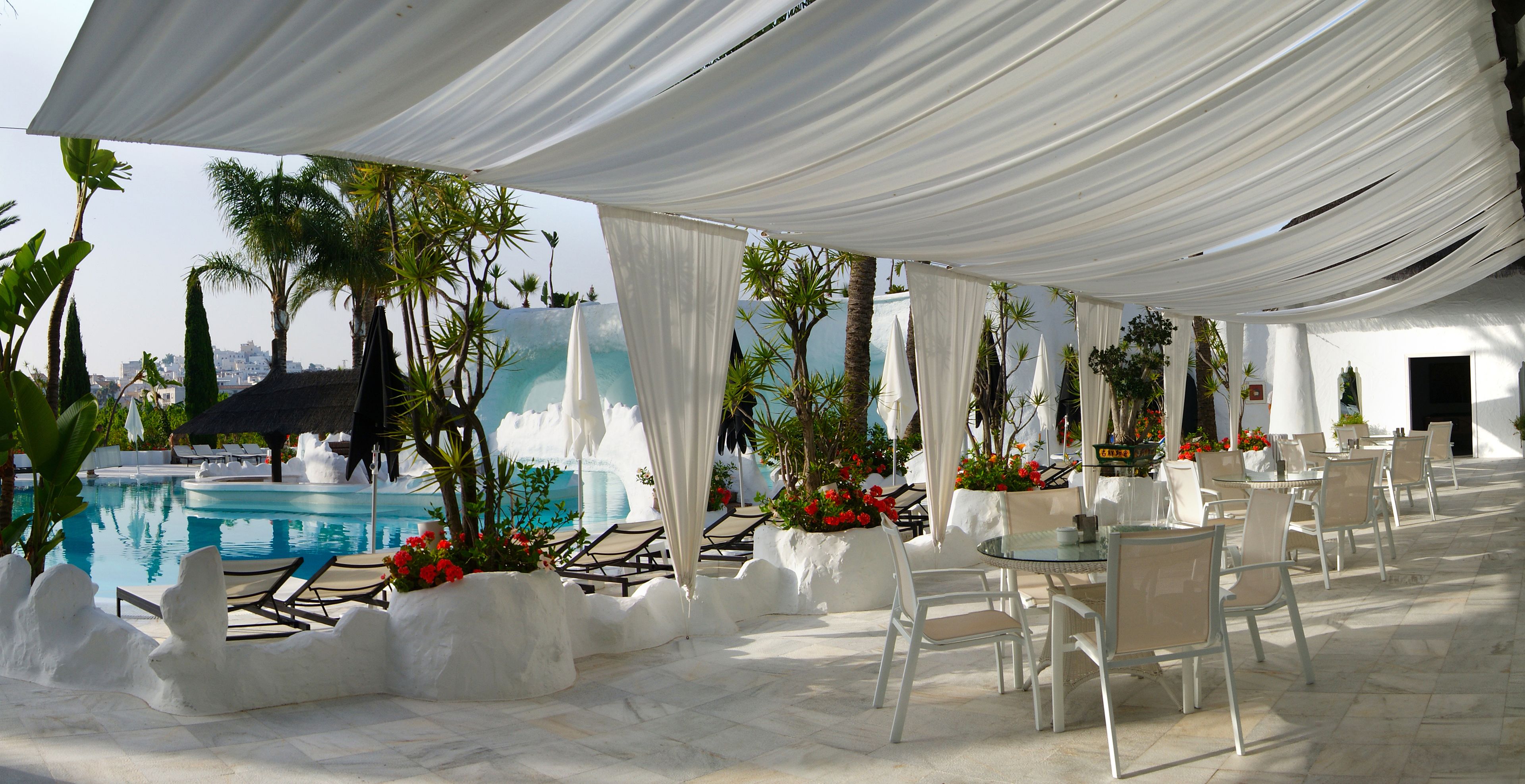 Un espace aménagé au bord de la piscine, avec des chaises longues, une table et un coin salon couvert agrémenté de rideaux blancs.
