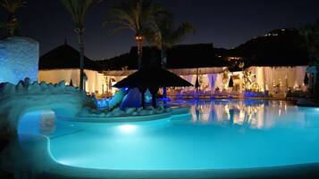 Un área de piscina nocturna con fuentes iluminadas, palmeras y una estructura de techo de paja.