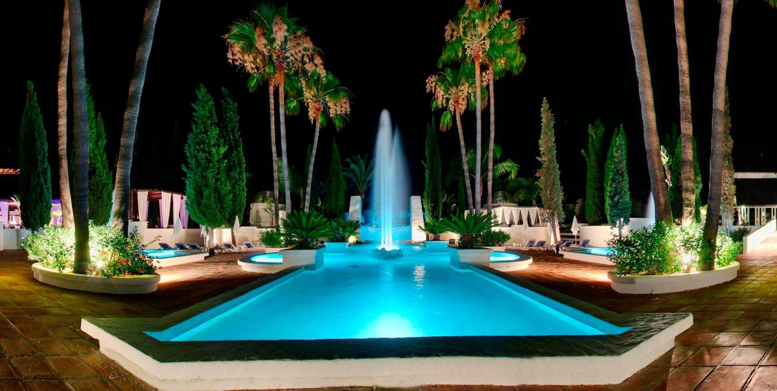 Una piscina iluminada de noche con una fuente central, rodeada de palmeras y sillas de descanso.
