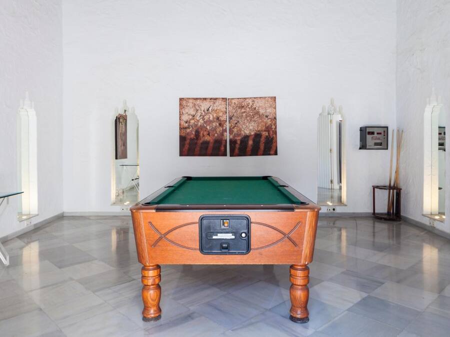Una mesa de billar en una habitación con suelo de baldosas, paredes blancas y un cuadro encima.