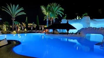 A nighttime swimming pool with palm trees and a thatched-roof structure.