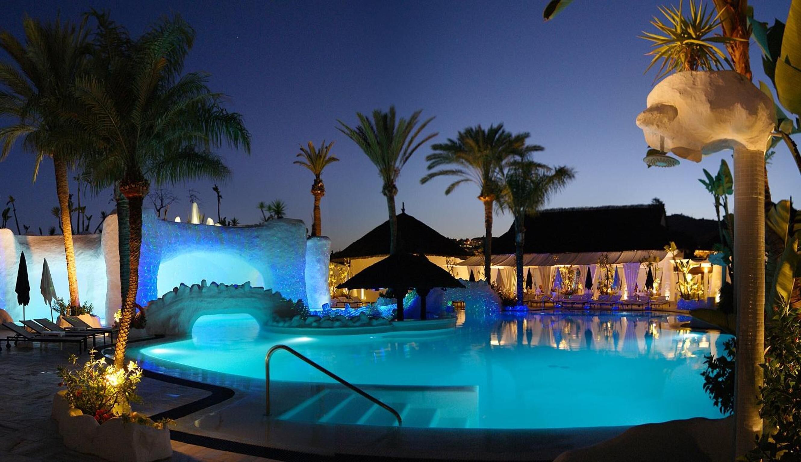 Area piscina notturna con vasca idromassaggio, palme e poltrone relax.