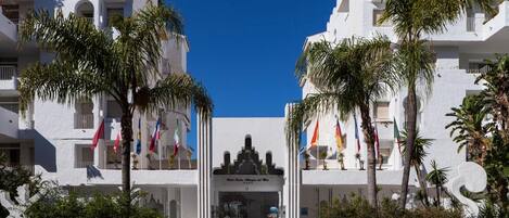 Un edificio blanco con balcones, palmeras, banderas y una entrada principal.