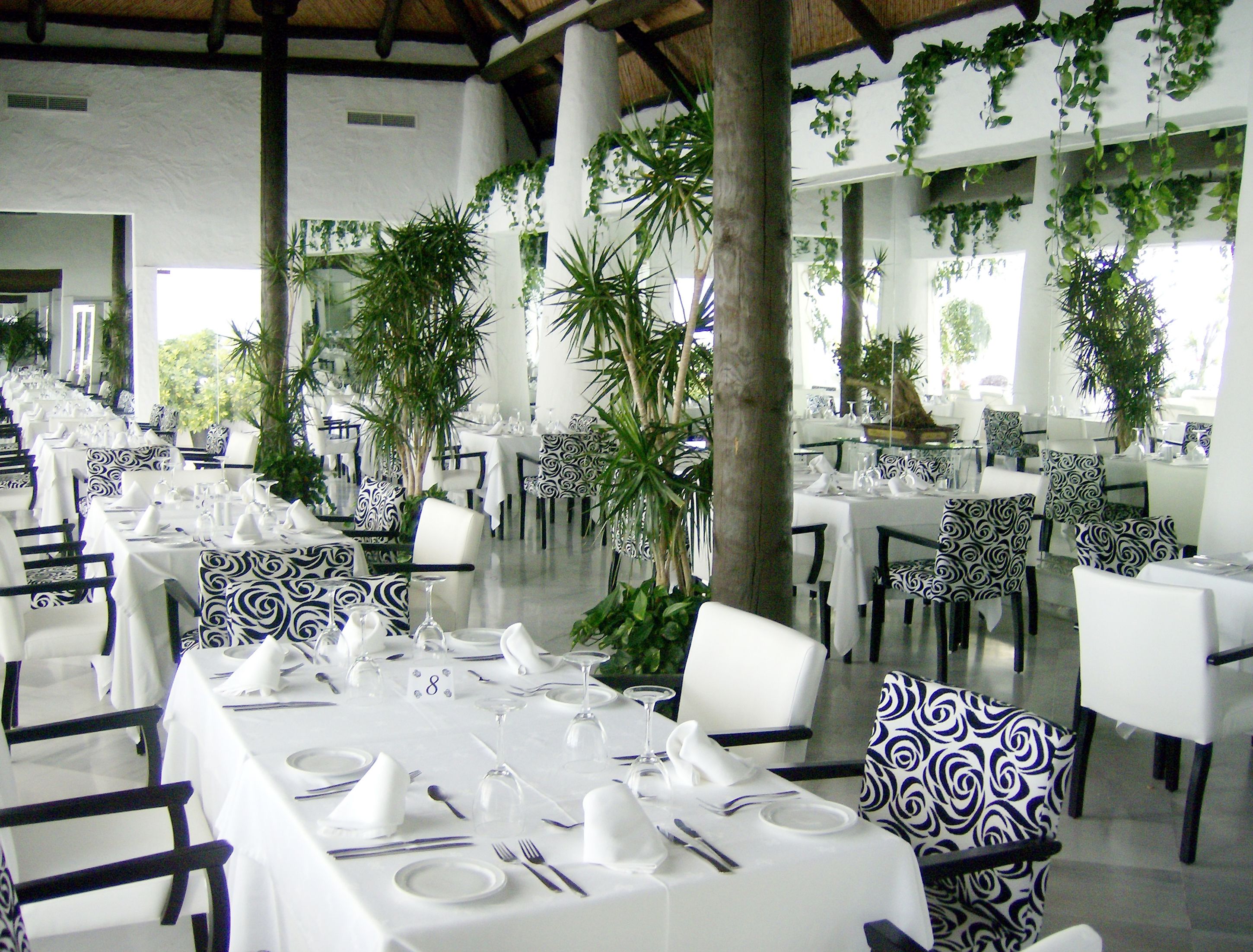 A restaurant with white tables and black and white patterned chairs, surrounded by greenery.