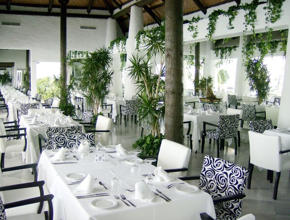 Un restaurante con mesas blancas y sillas con estampado blanco y negro, rodeado de vegetación.