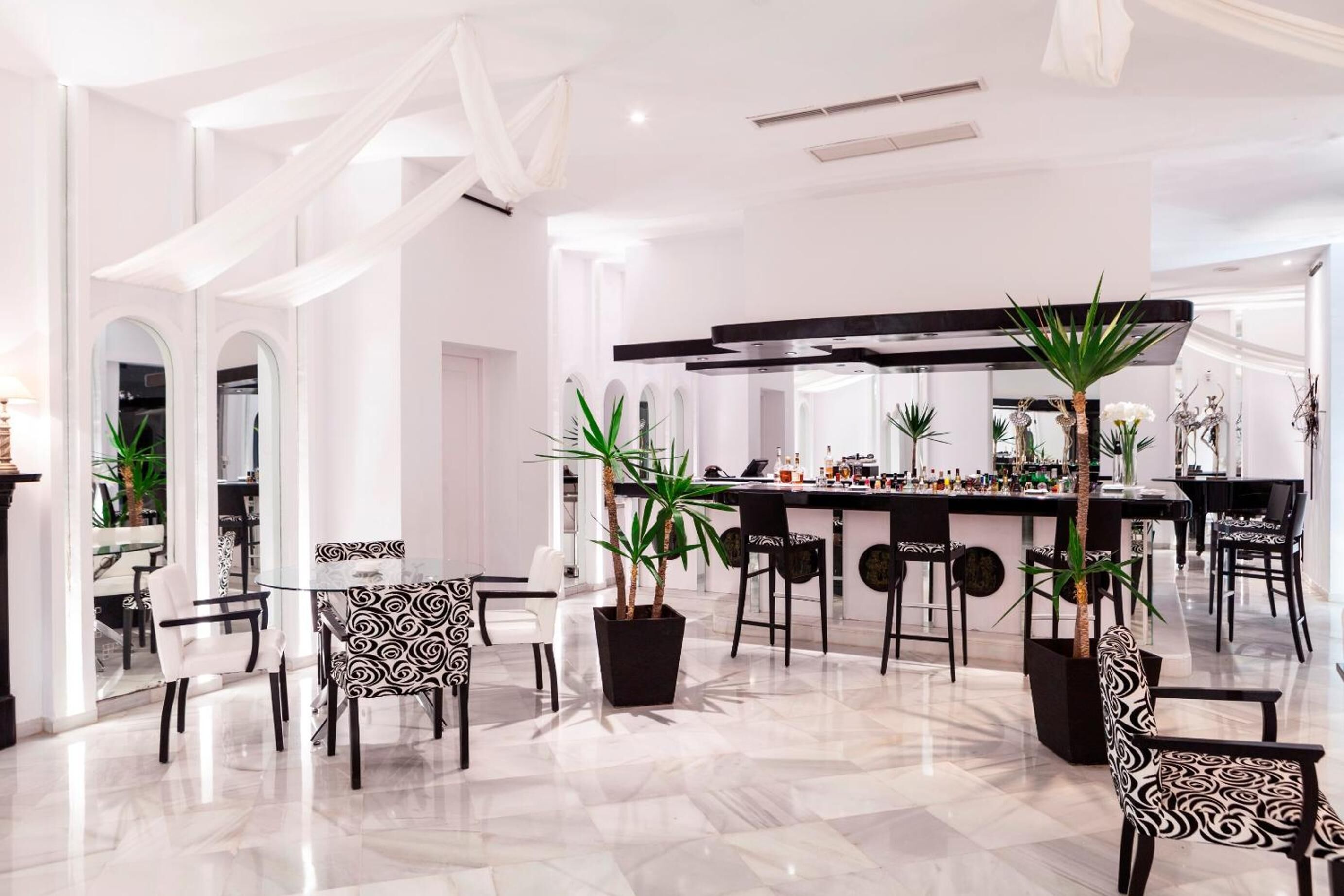 A modern dining area with a central bar, black and white patterned chairs, and potted plants.