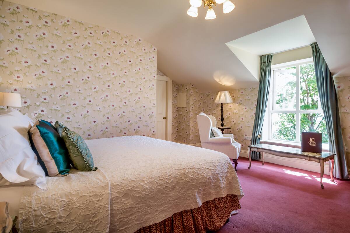 A bedroom with a large bed, a chair, a table, and a window with curtains.