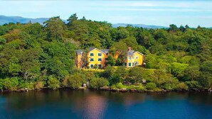 Una casa groga de dos pisos, amb un pràctic verd, envoltada per un bosc dens i una extensió d'aigua.