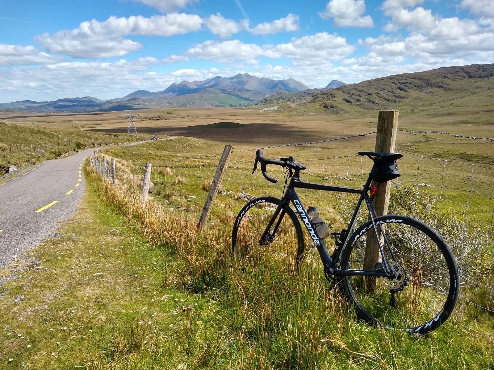 A bicycle parked by a fence on a rural road with mountains in the background.
