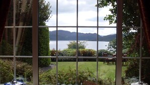 Finestra amb vista a un llac i muntanyes, jardí amb gespa i plantes en testos, i taula amb cadires a l'exterior.