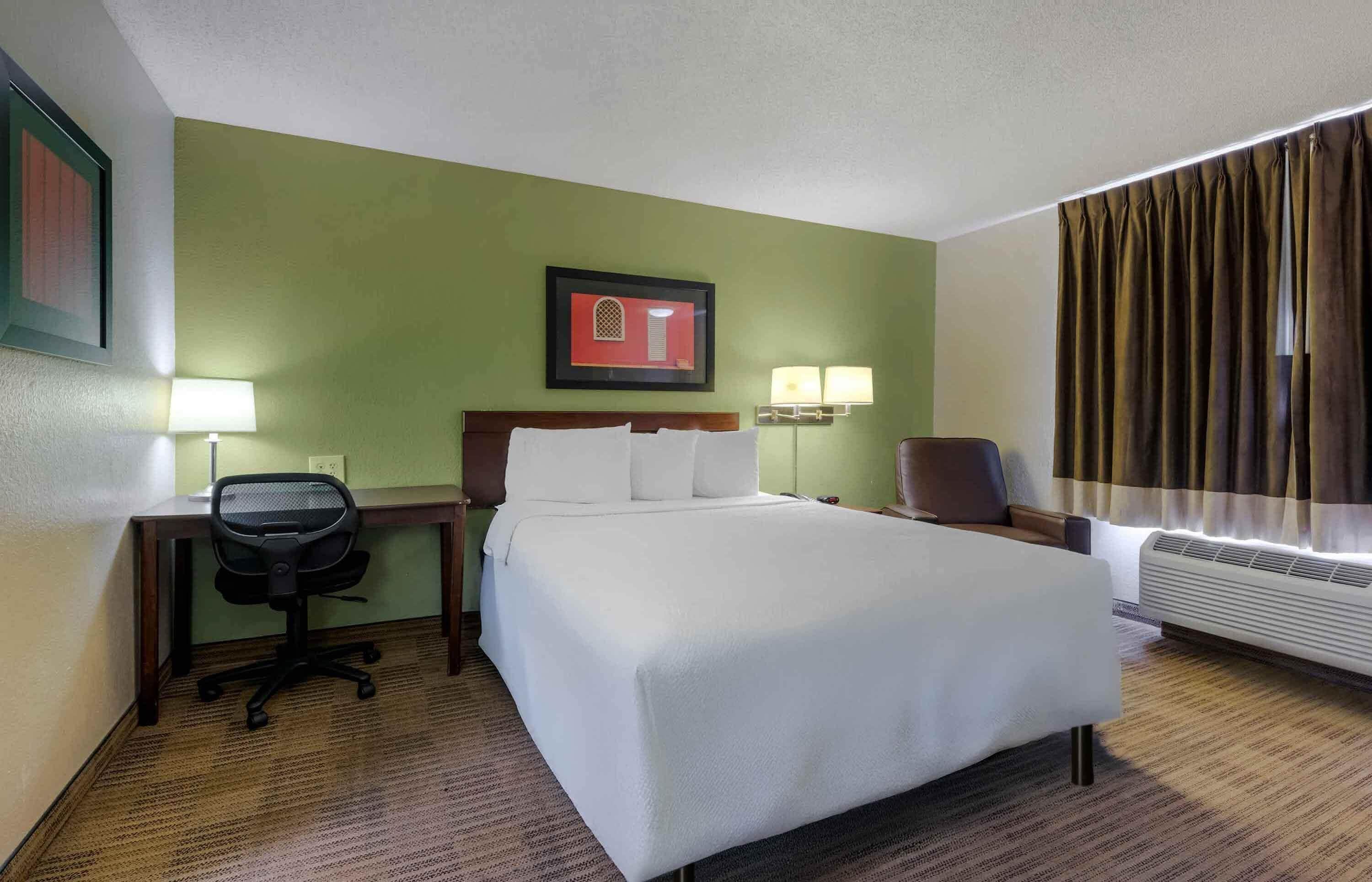 Quarto de hotel com cama, escrivaninha, cadeira, luminárias e janela com cortinas.