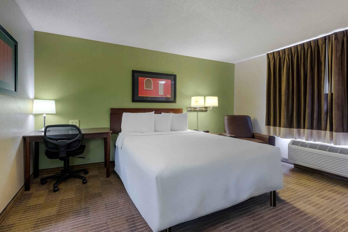 A hotel room with a bed, desk, chair, lamps, and a window with curtains.