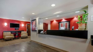 Un espace d’accueil avec un mur rouge, un comptoir de réception noir, deux fauteuils beiges, un téléviseur à écran plat et une plante en pot.