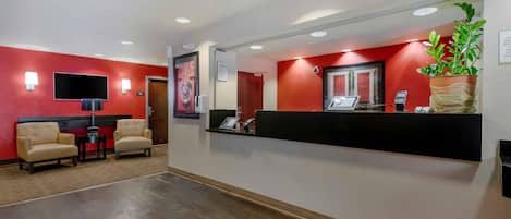 A reception area with a red wall, a black reception desk, two beige armchairs, a flat-screen TV, and a potted plant.