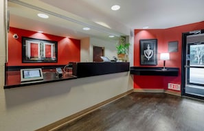 Un área de recepción con pared roja, mostrador negro, cartel de bienvenida enmarcado, planta en maceta y señal de salida.