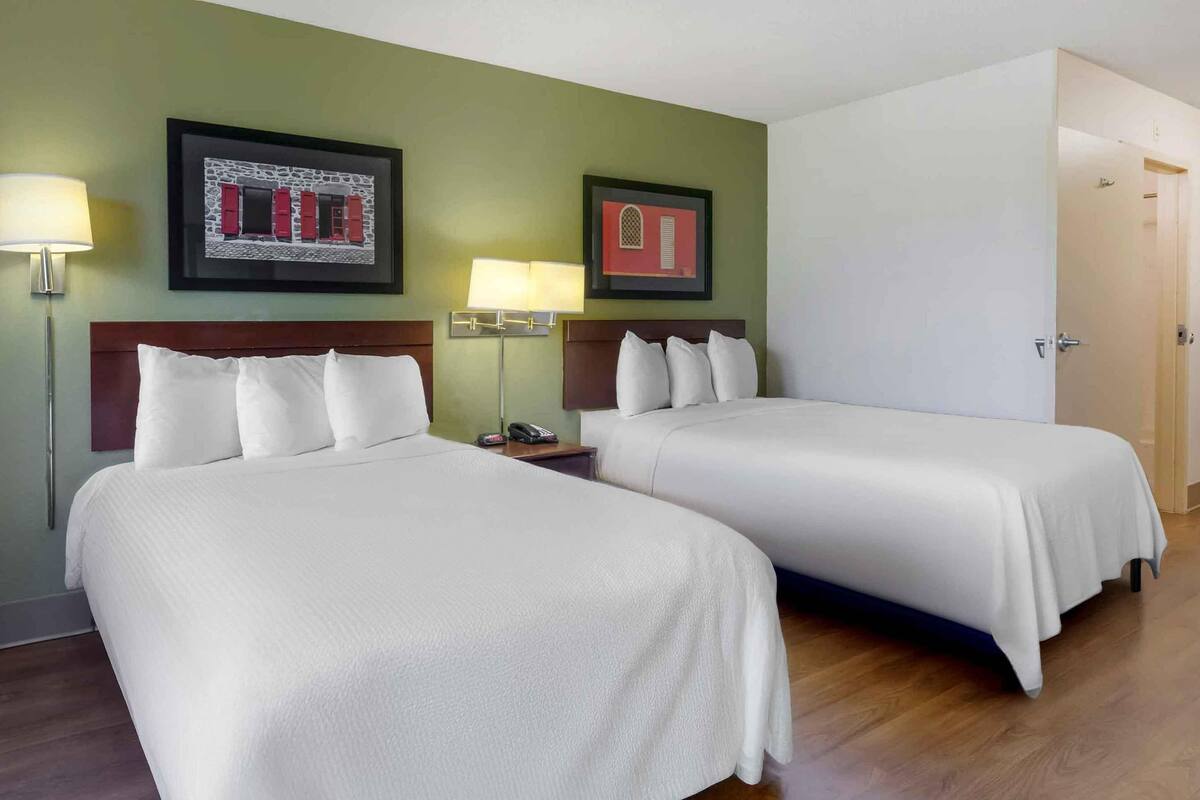 A hotel room with two beds, a wooden headboard, a nightstand with a lamp, and framed artwork on the wall.