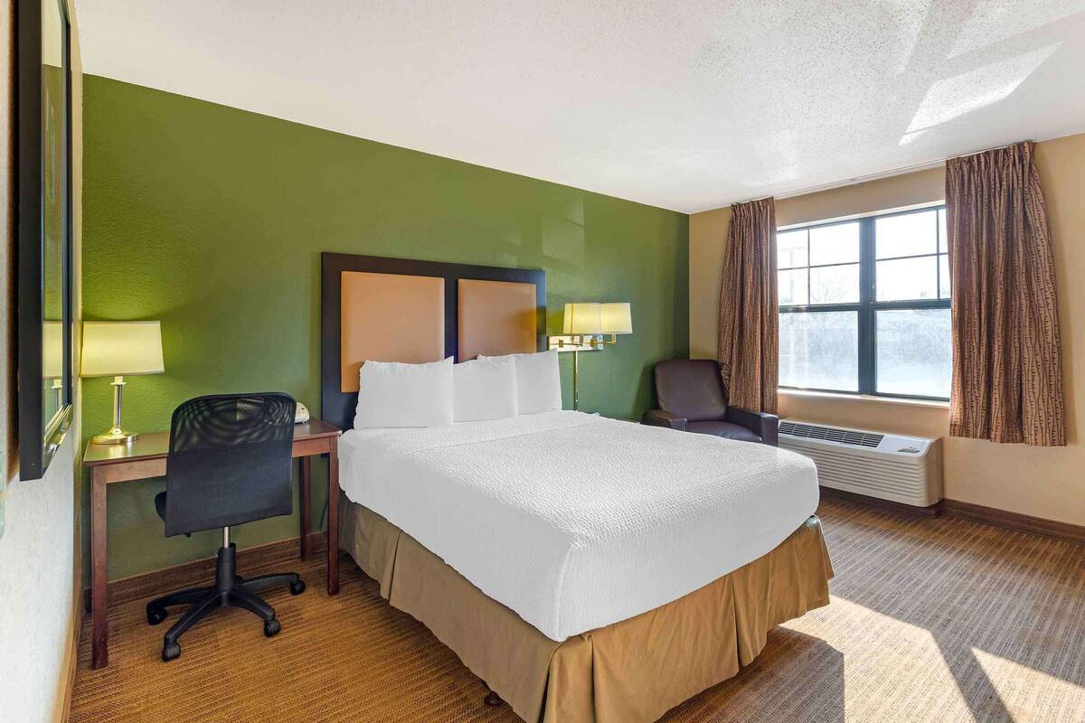 Une chambre d’hôtel avec un grand lit, un bureau avec une chaise, une lampe et une fenêtre avec des rideaux.