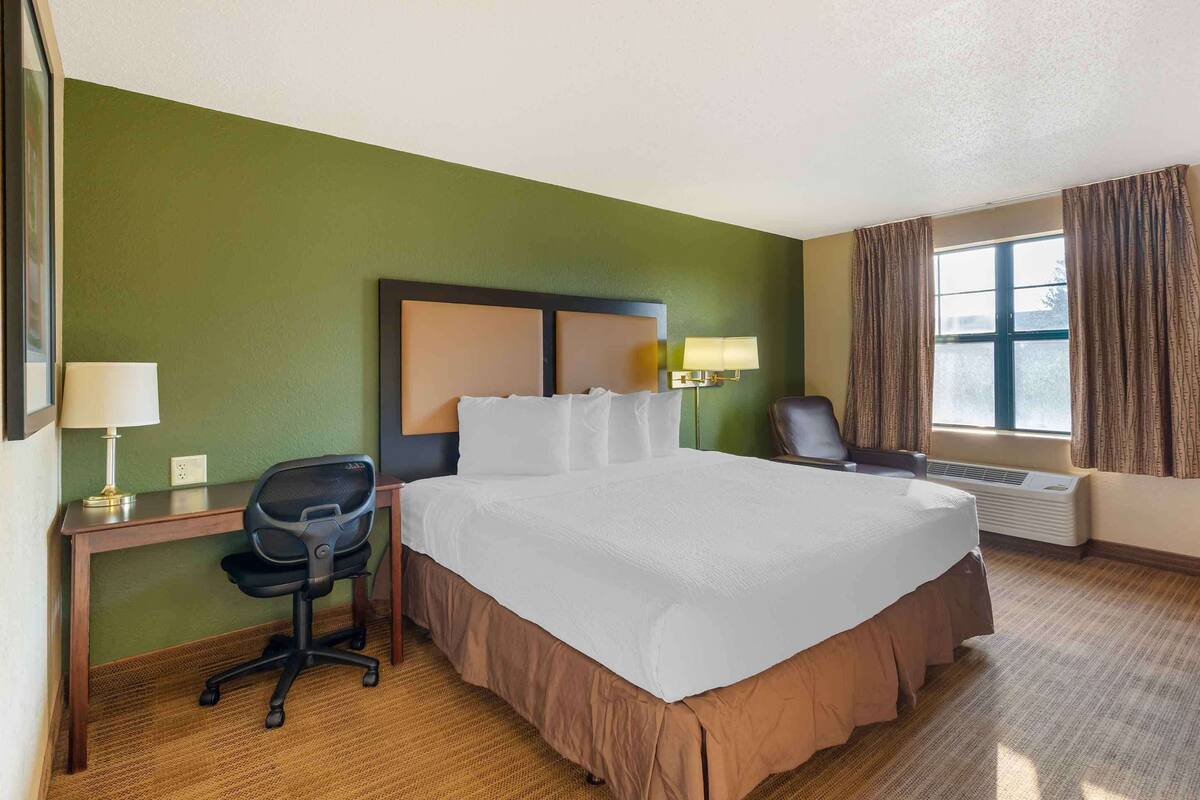 Une chambre d’hôtel avec un grand lit, un bureau avec une chaise, une lampe, une fenêtre avec des rideaux et un appareil de climatisation.