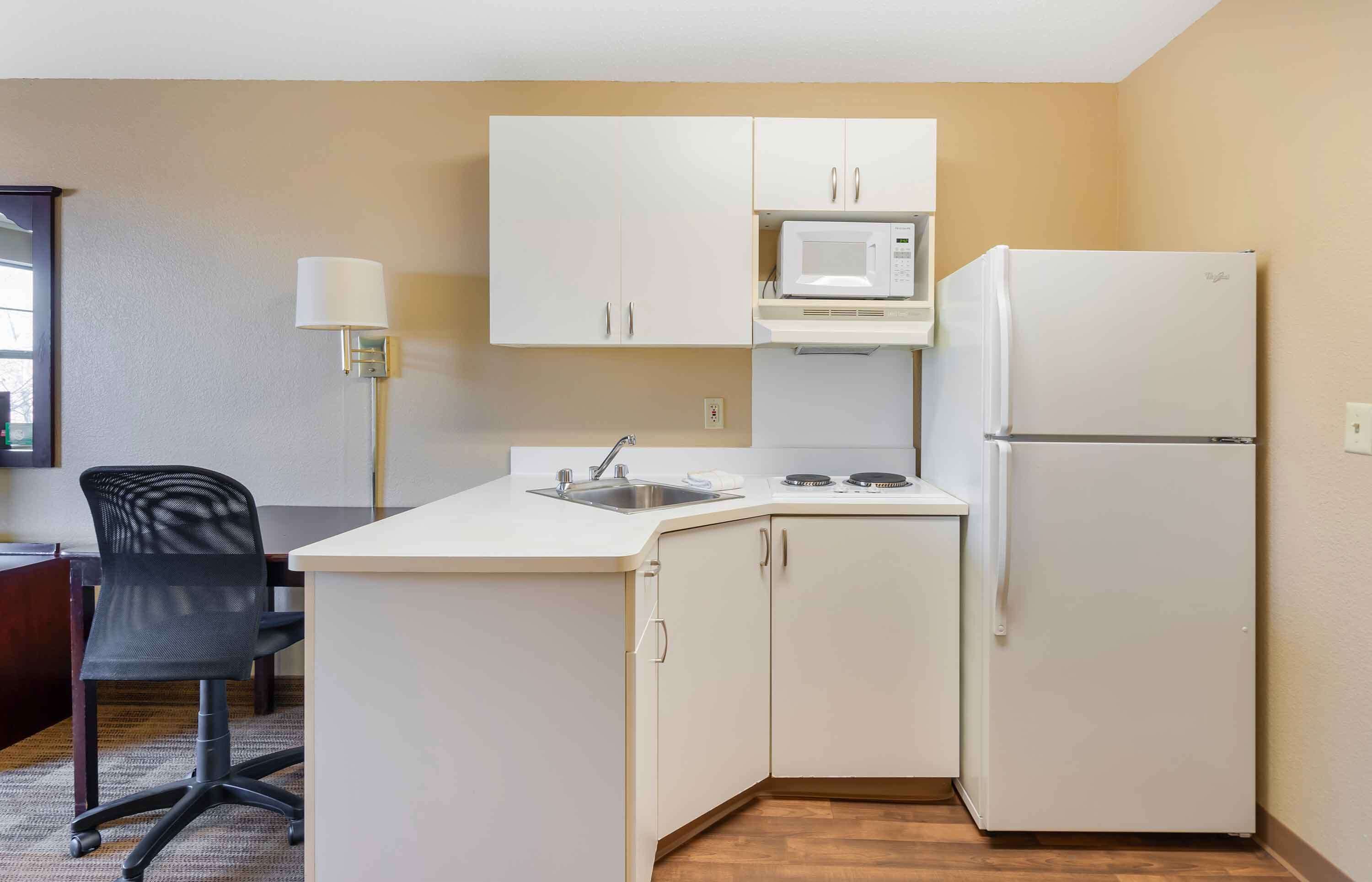 A compact kitchen with white cabinets, a microwave, a refrigerator, a sink, and a chair.