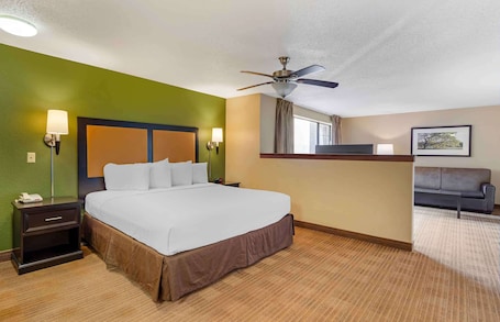 A hotel room with a bed, nightstands, a desk, a ceiling fan, a sofa, and a window with curtains.