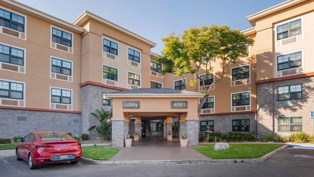 A multi-story hotel with a beige facade, a lobby entrance, and a red car parked in front.