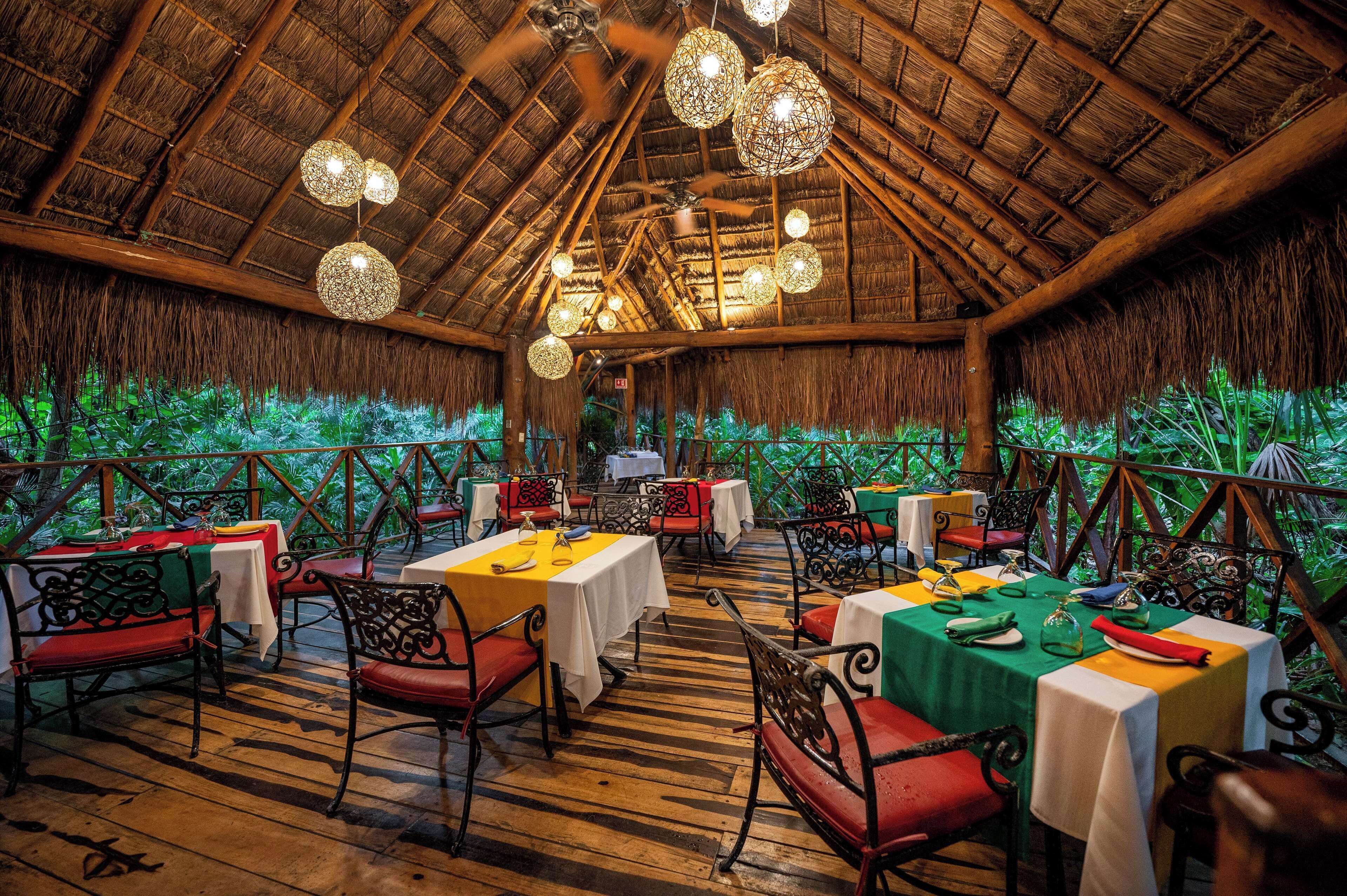 An indoor dining area with tables set for guests, a thatched roof, and hanging spherical light fixtures.