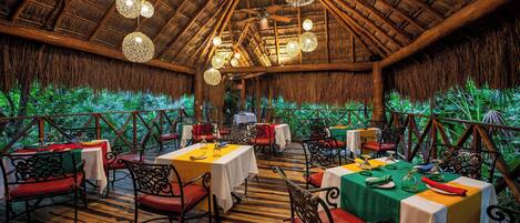 An indoor dining area with tables set for guests, a thatched roof, and hanging spherical light fixtures.