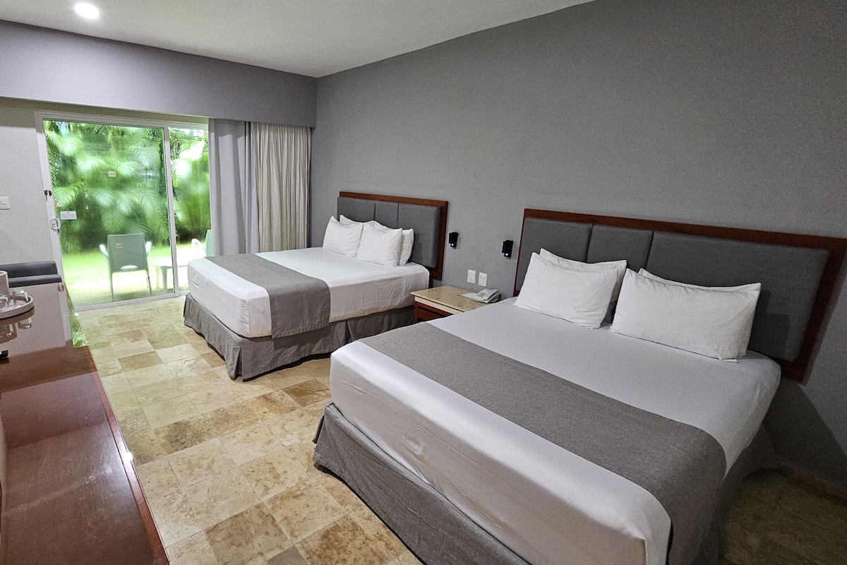 Habitación de hotel con dos camas, un escritorio y vistas a zonas verdes a través de un ventanal corredera.