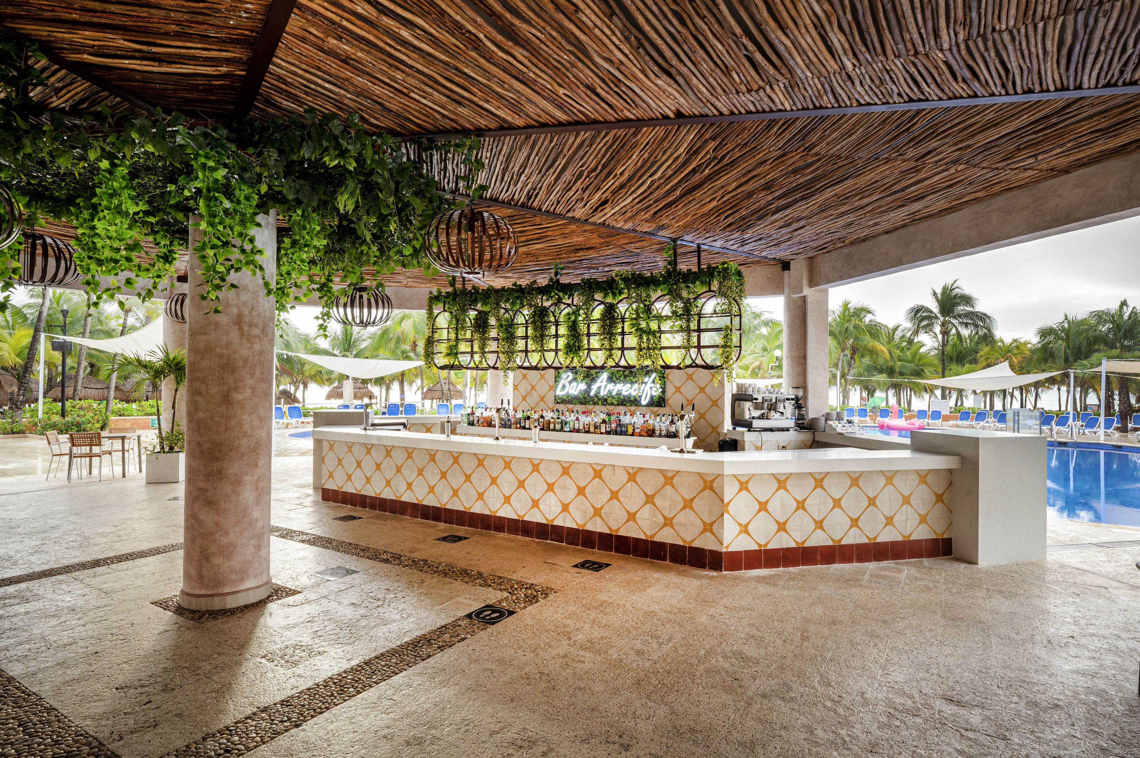 A bar area with a patterned counter, hanging plants, and a pool in the background.