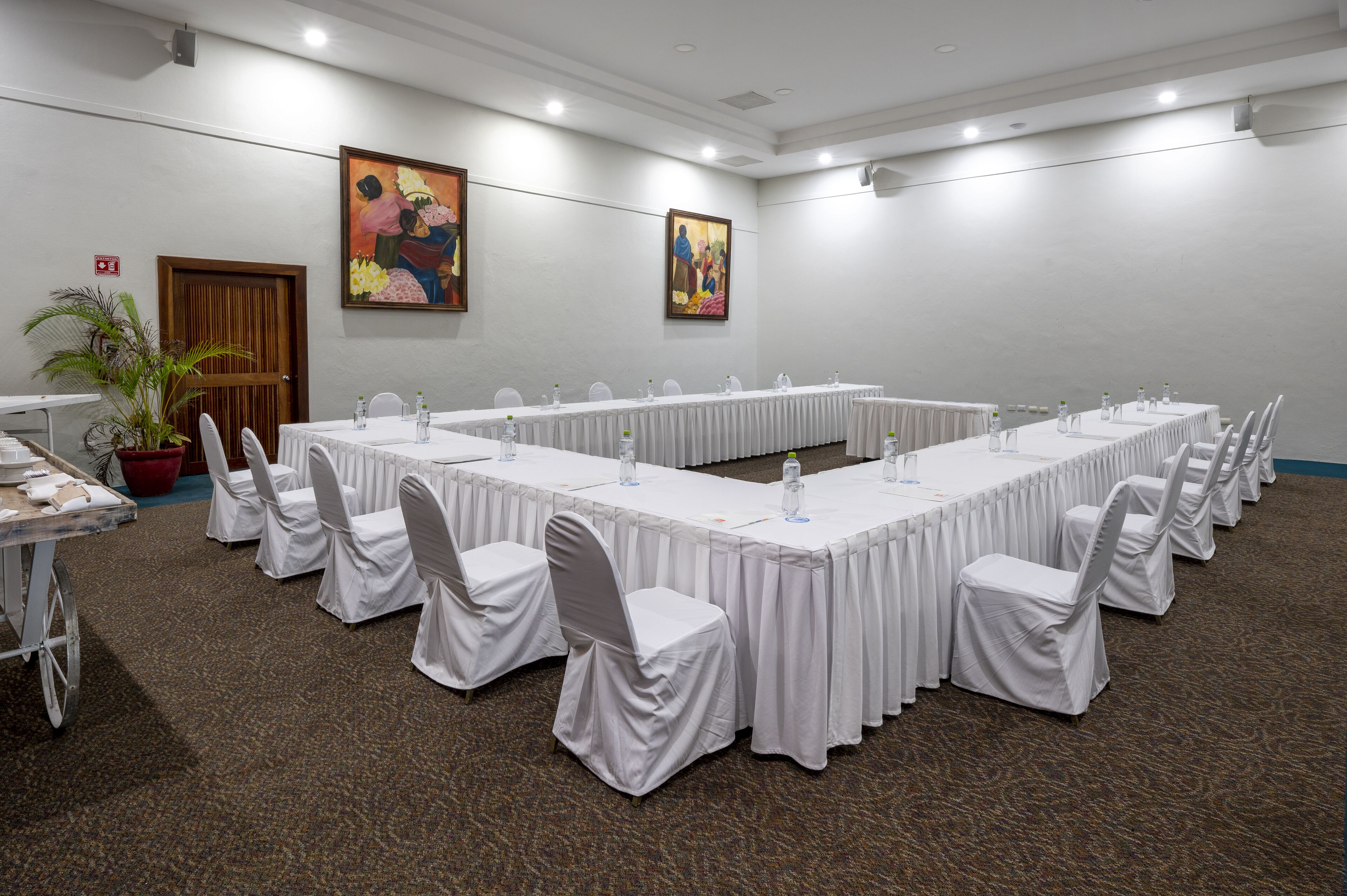 Un salón de conferencias con una larga mesa, sillas blancas y dos cuadros en la pared.