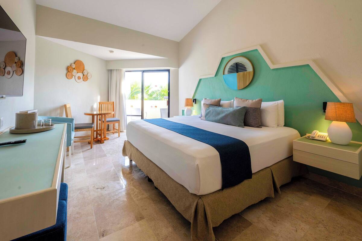 Habitación de hotel con una cama grande, un escritorio con una silla, una mesita y vistas al exterior a través de una puerta corrediza.