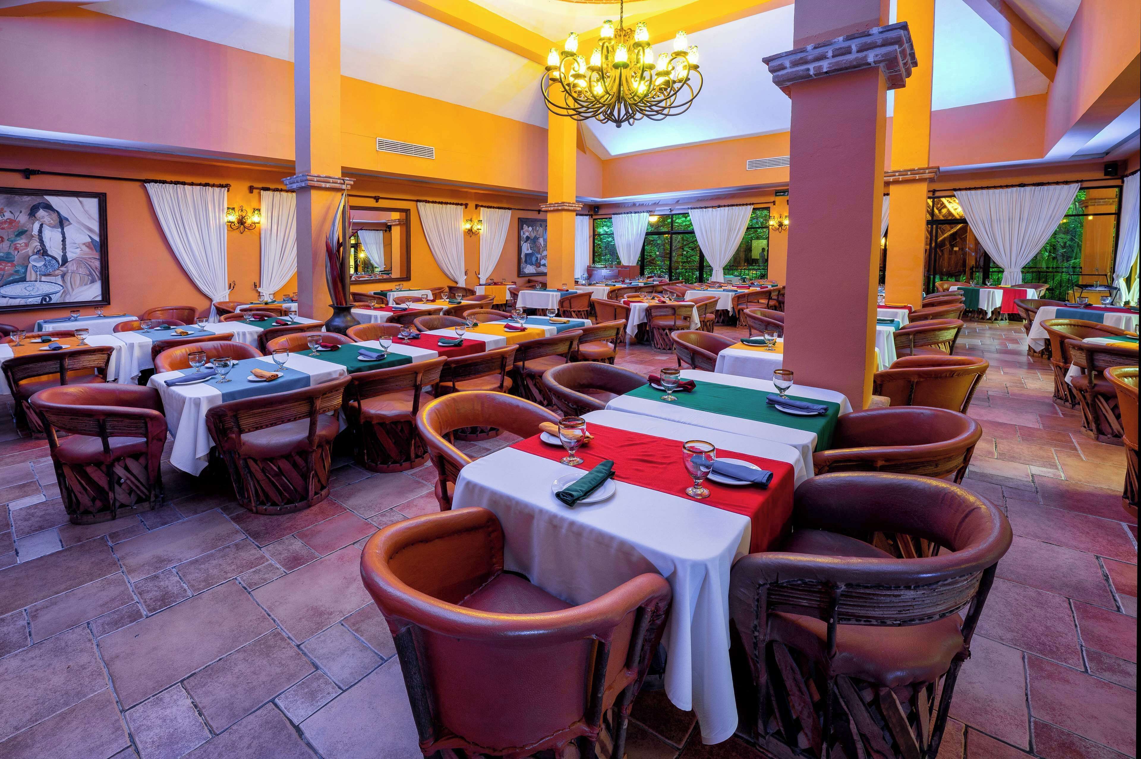 Un restaurante con varias mesas y sillas, una araña y ventanas amplias con cortinas.