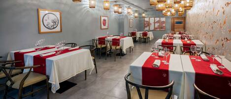 A restaurant with tables set for dining, featuring red and white tablecloths and wicker pendant lighting.