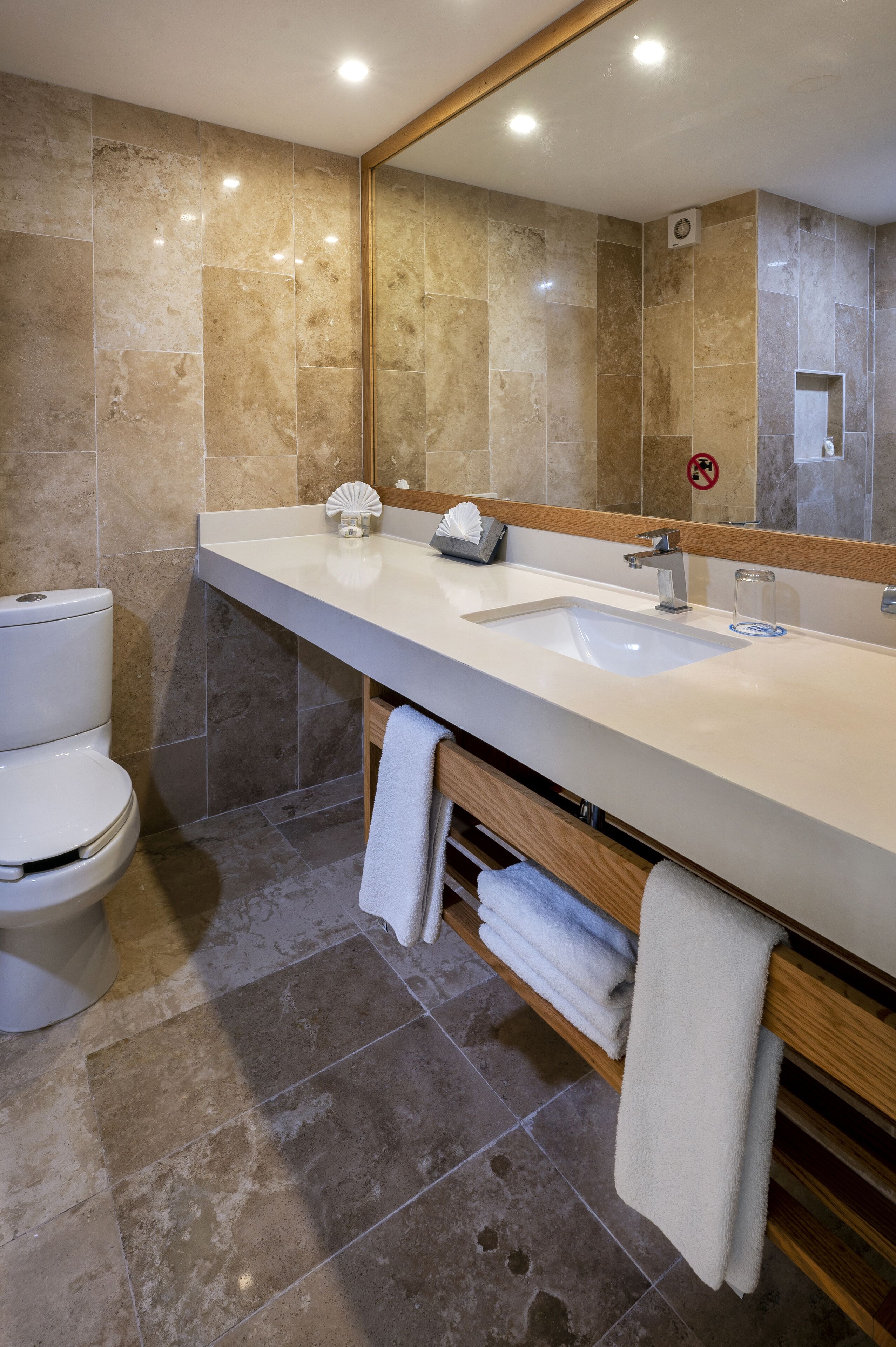 Un baño moderno con un revestimiento de baldosas color beige, un lavamanos blanco y un espejo sobre él.