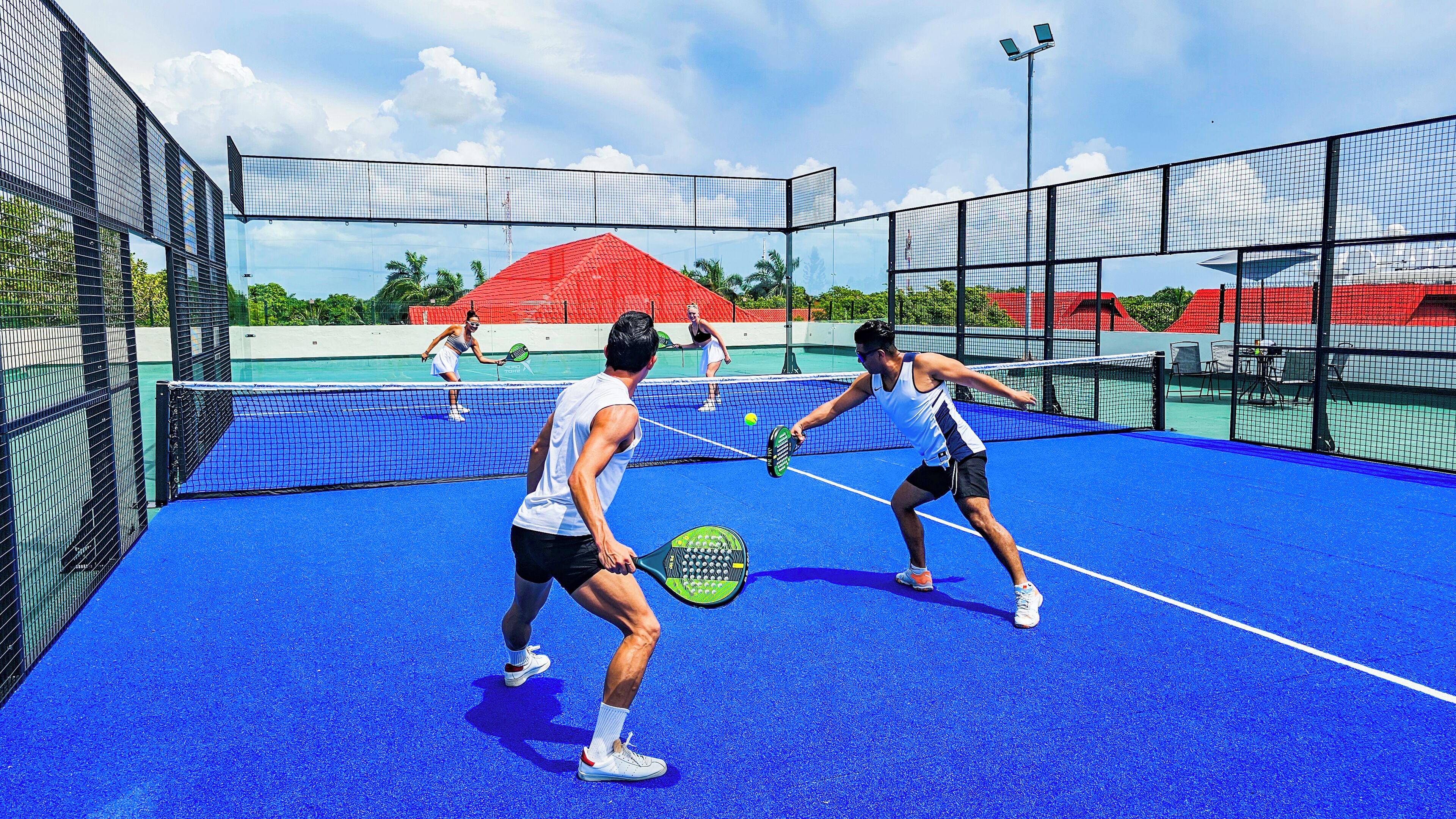 A tennis court with a blue surface, surrounded by a black fence, and a red-roofed building in the background.