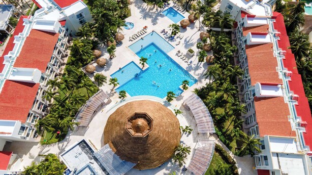 Eine Vogelperspektive zeigt einen Urlaubsort mit einem zentralen Swimmingpool, Palmen und einer kreisförmigen Struktur.